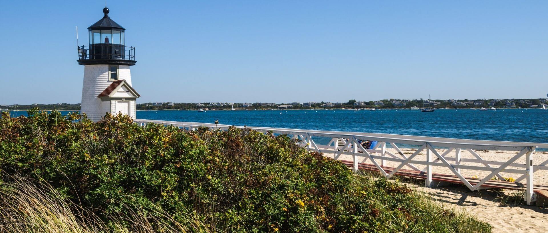 White lighthouse beside a wooden walkway overlooking blue water and boats in Nantucket Harbor.
