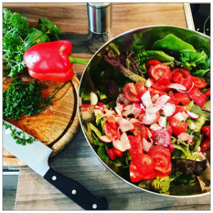 A bowl of bright green salad and vegetables in a bowl and a cutting board with a knife and a green pepper