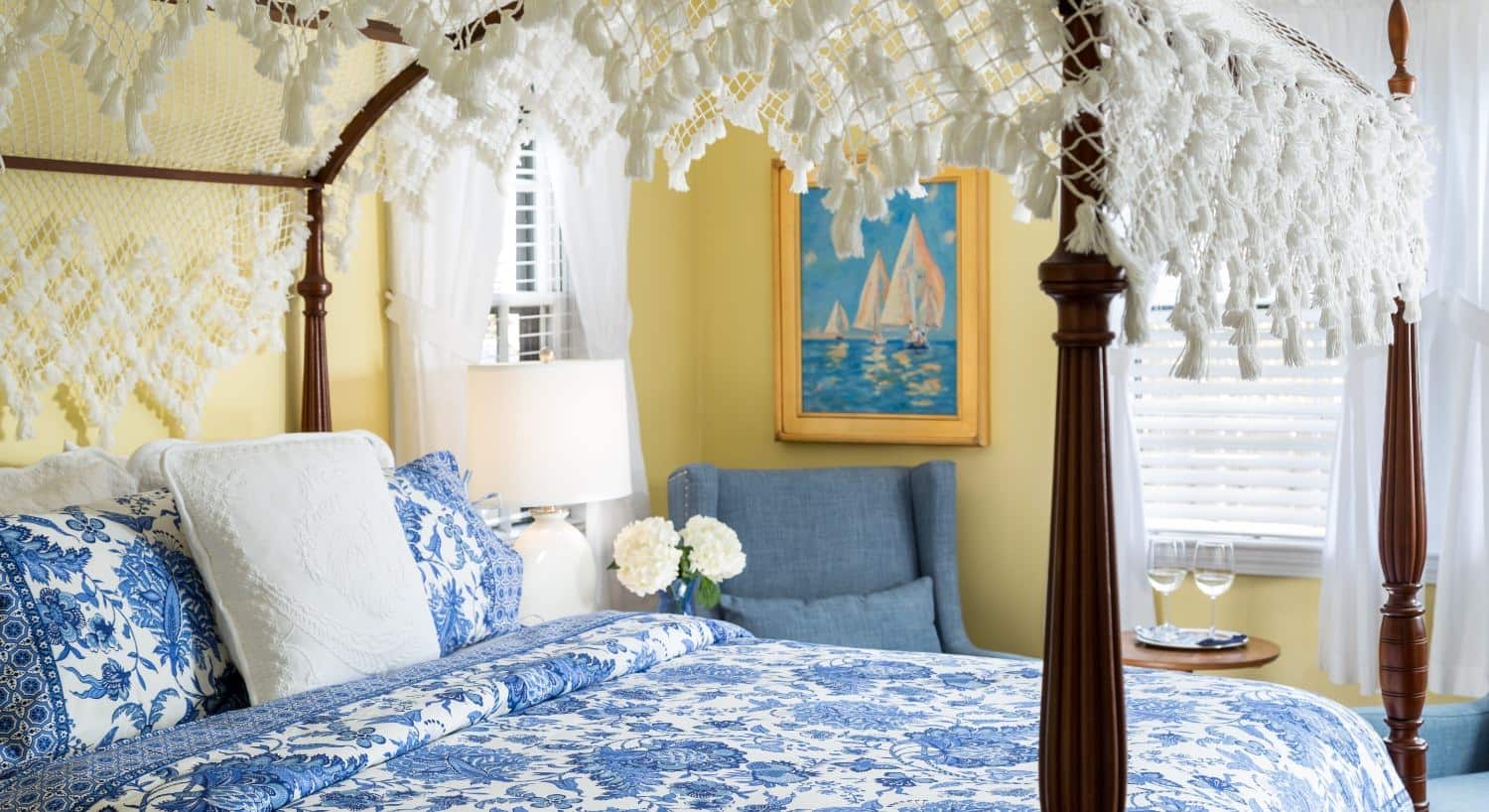 Bedroom with yellow walls, dark wooden four-poster bed, blue and white paisley bedding, denim upholstered arm chairs
