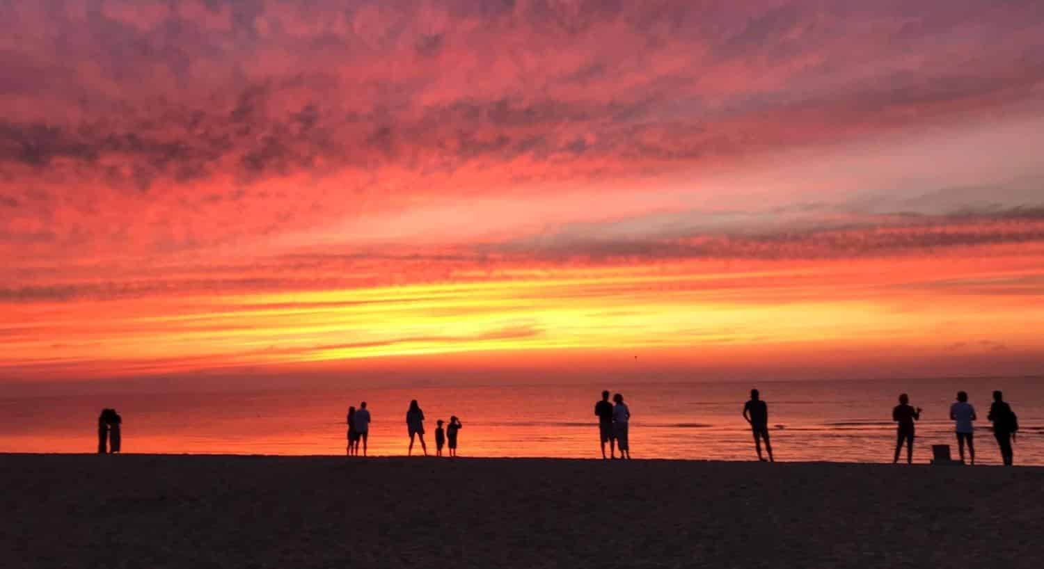 People on a beach watching a sunrise with clouds in the sky coloroed yellow, orange, pink, and purple
