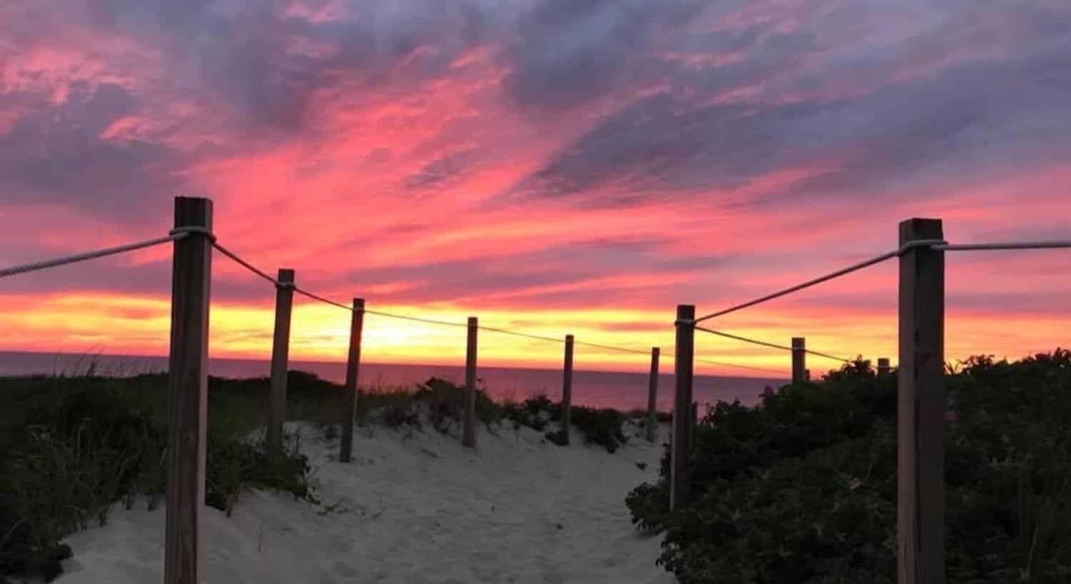 Sandy path with wooden posts and ropes leading to the beach with the sun rising in the background