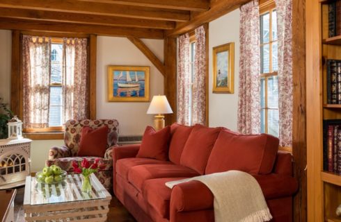 Living room with antique wooden fireplace, burgundy sofa, upholstered oversized armchair, and white wooden coffee table with glass top