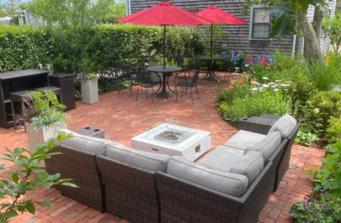 Exterior view of the property with large red brick patio with patio tables and chairs, couch around fire pit with blooming flowers, green privacy shrubs