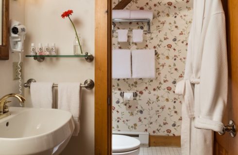 Bathroom with white pedestal sink, mirror, glass shelf, hanging white robe, and separate area with toilet