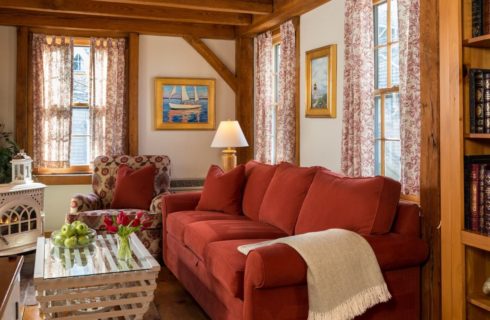 Living room with antique wooden fireplace, burgundy sofa, upholstered oversized armchair, and white wooden coffee table with glass top