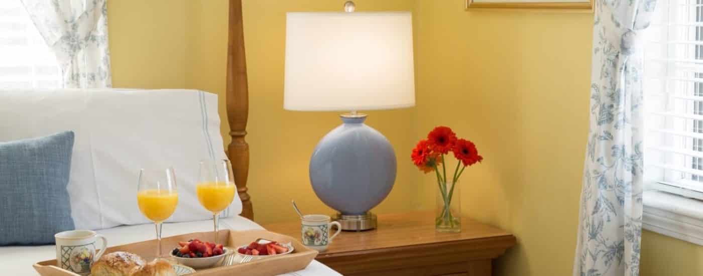 Bedroom with yellow walls, blue lamp with white shade on a light wooden nightstand, and tray full of pastries, fruit, juice, and coffee on top of the bed