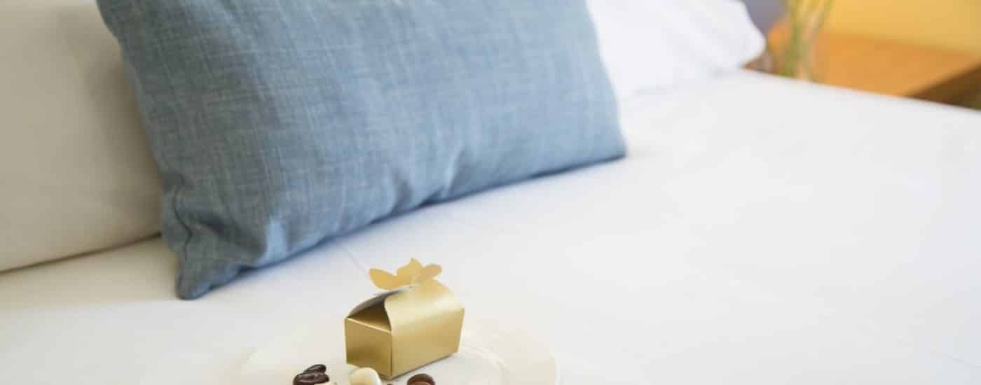 White plate full of chocolate candies and a gold box sitting on top of a bed with white bedding and blue denim pillow