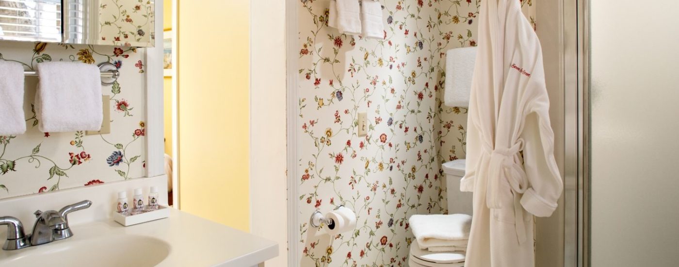 Bathroom with multicolored floral wall paper, white wooden vanity, stand up shower, and hanging white robe