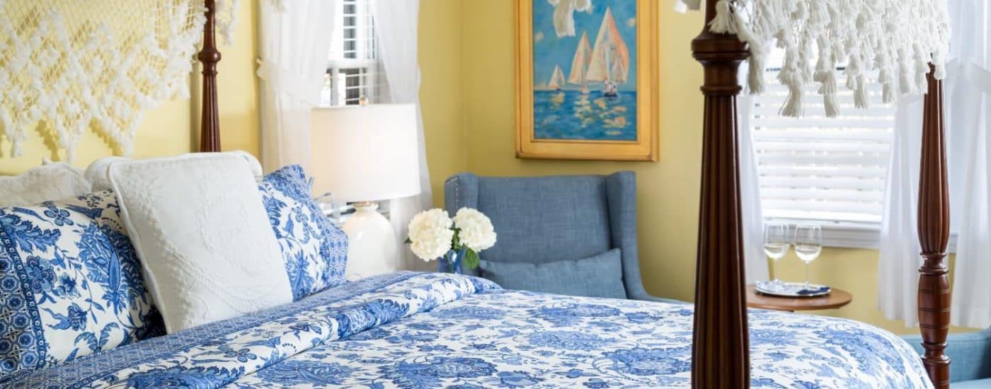 Bedroom with yellow walls, dark wooden four-poster bed, blue and white paisley bedding, denim upholstered arm chairs