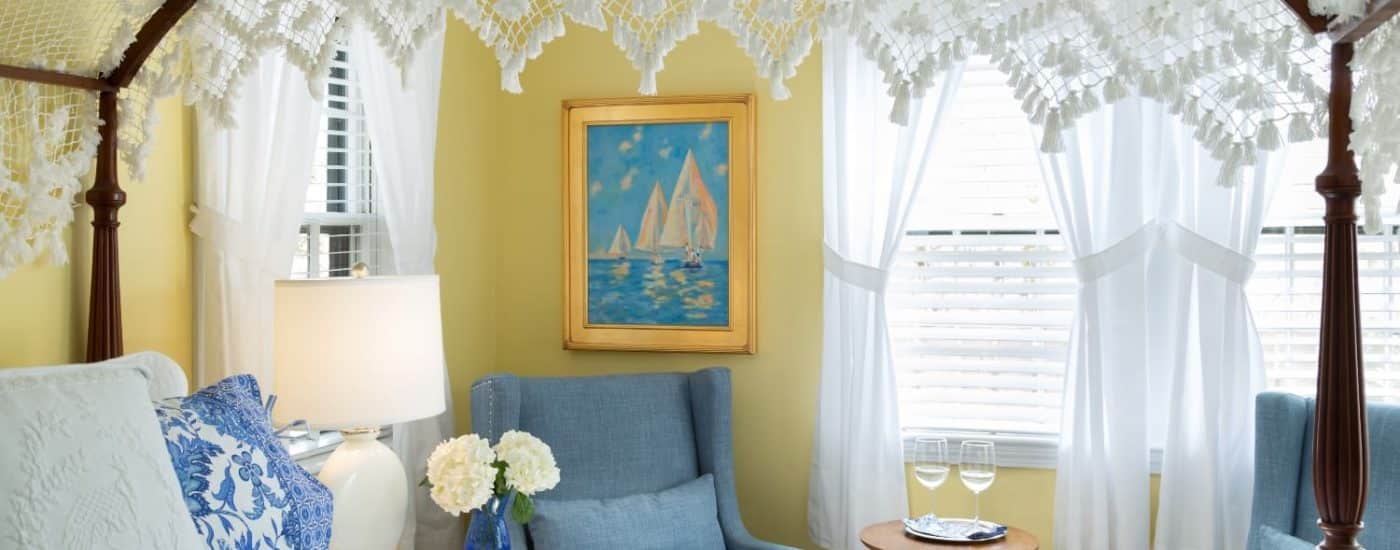 Bedroom with yellow walls, dark wooden four-poster bed, blue and white paisley bedding, denim upholstered arm chairs