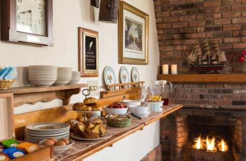 Dining area with a wooden counter filled with continental breakfast items next to a large brick fireplace