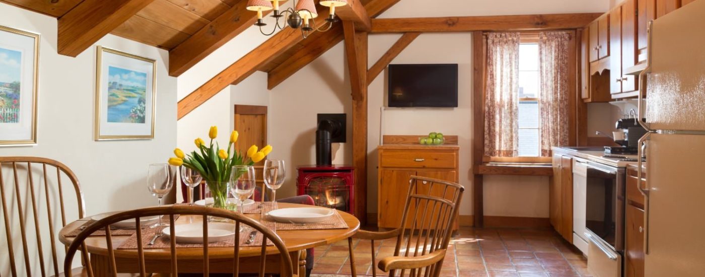 Kitchen with white walls and terra cotta tile flooring, wooden table, chairs, and cabinets, refrigerator, stove, dishwasher, flat-screen TV, and fireplace