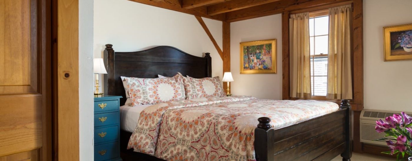 Bedroom with dark wooden bed, multicolored bedding, teal-colored wooden dresser, wood trim around window, and exposed wood ceiling joists