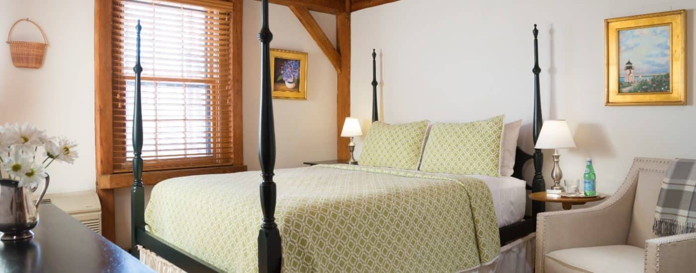 Bedroom with white walls, wood window frame, dark wooden four-poster bed, and upholstered arm chair