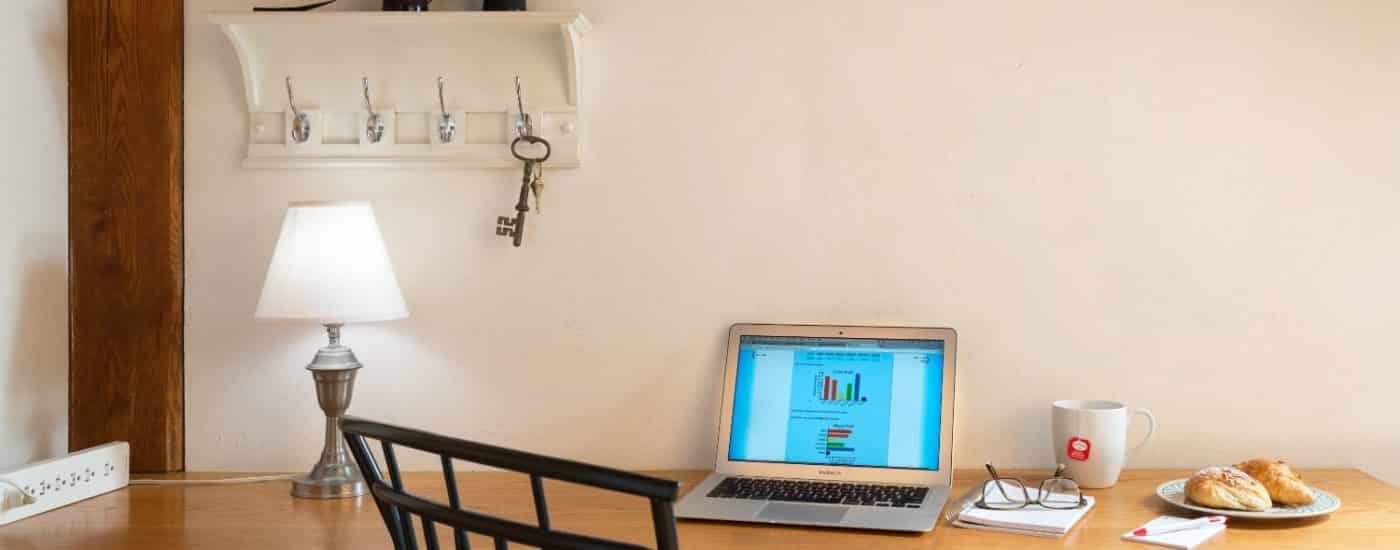 Large wooden desk with a powerstrip, desk lamp, laptop, mug, notepad, and a small plate with pastries