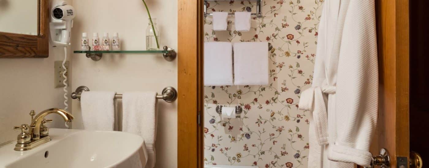 Bathroom with white pedestal sink, mirror, glass shelf, hanging white robe, and separate area with toilet