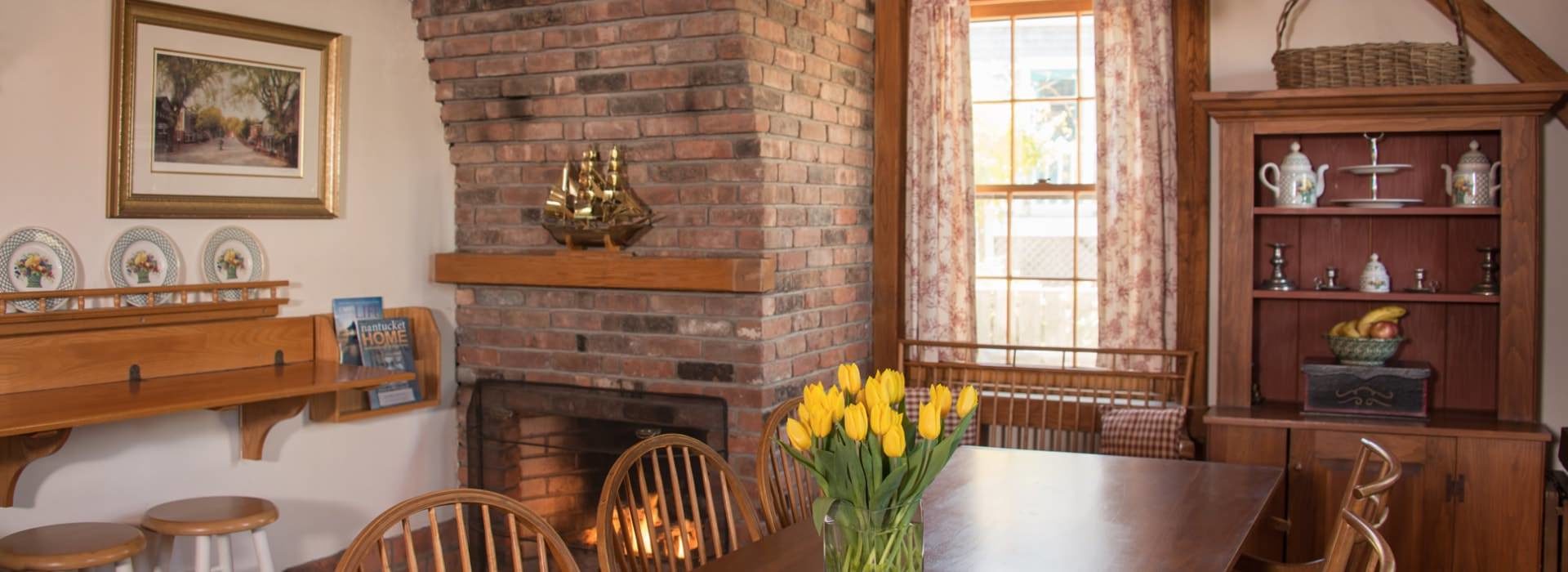 Dining room with large brick fireplace and large wooden table and chairs with yellow tulips in a vase on top