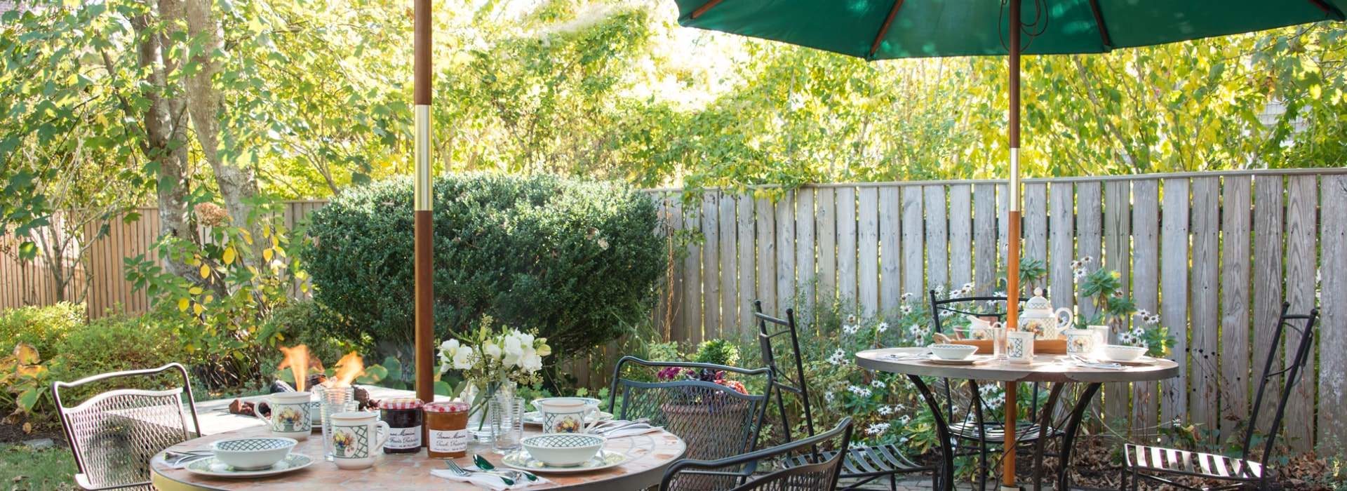 Two patio tables with opened umbrellas and place settings on a patio with green shrubs, trees, and flowers