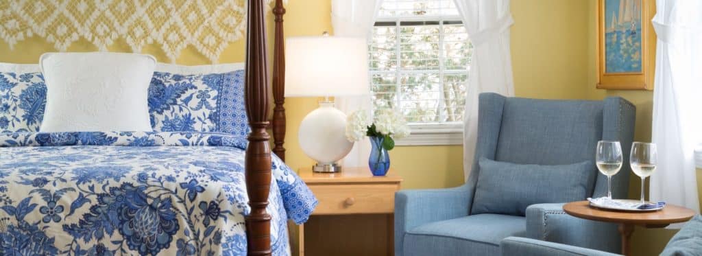 Bedroom with yellow walls, dark wooden four-poster bed, blue and white paisley bedding, denim upholstered arm chair
