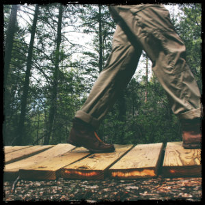 Man walking in hiking boots across a footbridge in a green wooded forest. Image by lacey-raper-361 www.unsplash.com