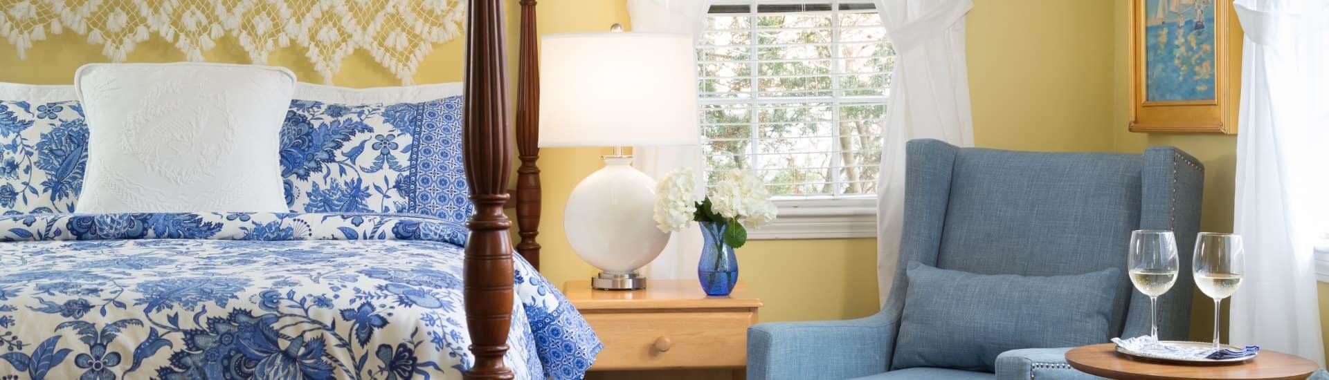 Bedroom with yellow walls, dark wooden four-poster bed, blue and white paisley bedding, denim upholstered arm chair