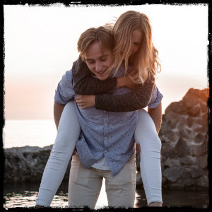 Man in light pants and blue shirt carrying a blonde woman wearing white pants and dark shirt on his back 