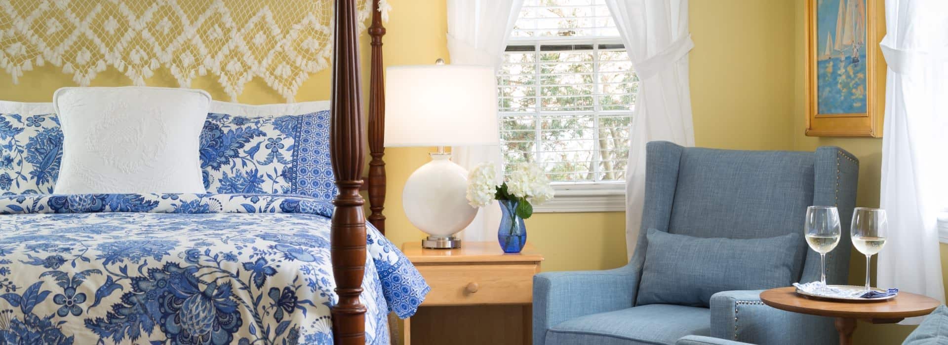 Bedroom with yellow walls, dark wooden four-poster bed, blue and white paisley bedding, denim upholstered arm chair