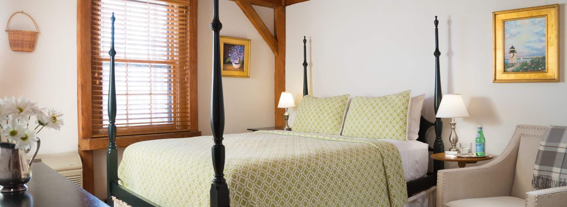 Bedroom with white walls, wood window frame, dark wooden four-poster bed, and upholstered arm chair